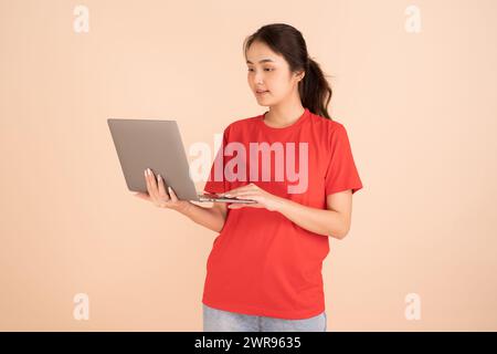 A beautiful Asian woman in a red shirt is holding a laptop computer on a cream background She is having fun surfing the web. Stock Photo