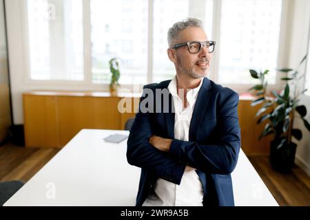 A mature professional stands with arms crossed, a slight smile on his face, in a light-filled office, suggesting both experience and approachability Stock Photo