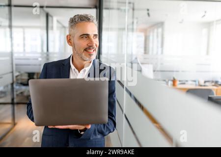 Purposeful mature businessman in smart casual wear stand in modern co-working space and holding laptop, male freelancer or start-up owner with laptop computer indoors looking away cheerfully Stock Photo