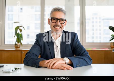 Portrait of an elegant mature businessman wearing glasses and smart casual jacket smiling at the camera. Webcam view during video call Stock Photo