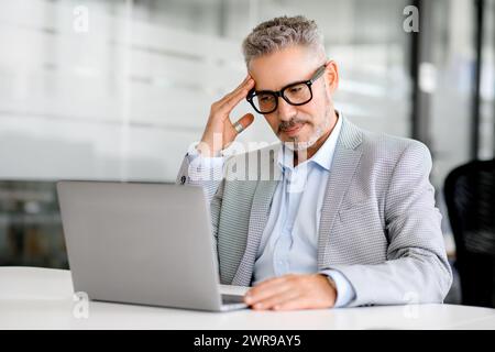 Mature businessman is deeply focused on his laptop screen in a serene office setting, embodying the concentration and strategic thought that drives business success, a moment of deep analysis Stock Photo