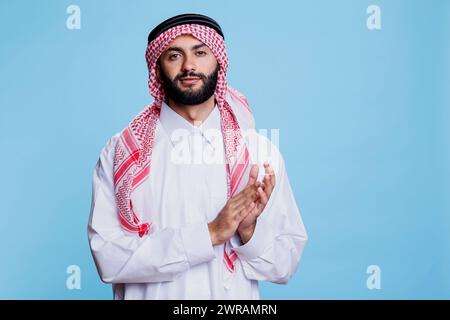 Confident muslim man wearing traditional islamic clothes applauding while looking at camera. Young arab dressed in white thobe and checkered ghutra clapping hands studio portrait Stock Photo
