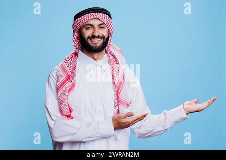 Muslim man in traditional robe confidently pointing to left side with hands studio portrait. Arab showcasing product advertisement and looking at camera with cheerful expression Stock Photo