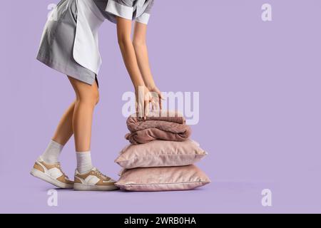 Female housekeeper with stack of pillows and warm plaid on lilac background Stock Photo