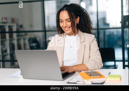 Amazing curly haired hispanic or brazilian young woman in stylish elegant clothes, office employee, small business owner, working on a laptop while sitting at her workplace in the modern office, smile Stock Photo