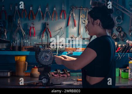 Mixed-race hispanic young Female mechanic, wear black overall, focused on an motorcycle part in a workshop with a tool cabinet, a complete tool panel Stock Photo