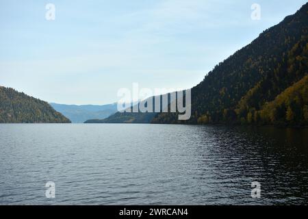 The wide and calm surface of a large lake surrounded by high gentle mountains covered with dense coniferous forest on a sunny autumn evening. Lake Tel Stock Photo