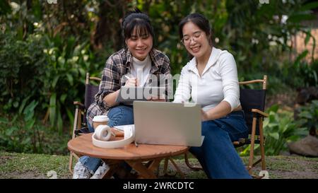 Two young Asian female friends, college students, are talking, looking at a laptop screen, and working on a project together while sitting at an outdo Stock Photo