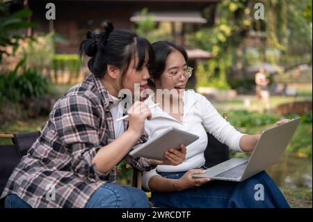 Two young Asian female friends, college students, are talking, looking at a laptop screen, and working on a project together while sitting at an outdo Stock Photo