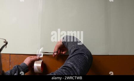 a man uses scissors to cut off a strip of tape glued at the junction of two polyester sheets of soft insulation on the floor, preparing and fixing Stock Photo