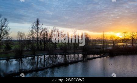 This image captures a serene dusk setting by a riverside, featuring the silhouettes of trees against a softly glowing sky. The sun, near the horizon, bathes the clouds in a gradient of orange and yellow, contrasting with the cooler blues of the impending night sky. The calm river mirrors the silhouettes, enhancing the quietude of the scene. The distant landscape is bathed in the sunset's fading light, providing depth and context to the rural environment. Dusk Silhouettes and Reflections by the Riverside. High quality photo Stock Photo