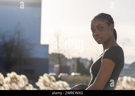 The photograph shows a pensive African American woman outdoors at sunset. Her braided hair drapes over her shoulders, and she wears a dark shirt, merging with the early evening light. The blurred urban environment in the background, with its buildings and clear sky, creates a contemplative atmosphere. Contemplative Young Woman in Urban Sunset. High quality photo Stock Photo