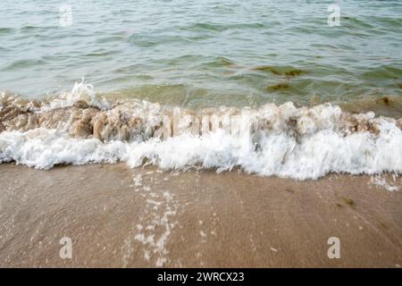 The image captures the dynamic interaction between sea and shore as a wave crashes onto the sandy beach. The frothy white of the wave contrasts with the murky green of the coastal waters, illustrating the perpetual motion of the ocean. Sand particles are visible in the clear water, stirred up by the force of the wave, while the foam bubbles up, adding texture to the scene. The photograph conveys the continuous cycle of waves that sculpt and define the coastline, a natural rhythm that is both powerful and calming to witness. Dynamic Seashore: Waves Crashing on Sandy Beach. High quality photo Stock Photo