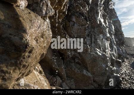 This image offers a detailed close-up of water cascading down a rough, textured rock face on a sunny beach. The sunlight catches individual droplets, highlighting their clarity and the intricate paths they follow down the stone surface. The perspective is up-close, emphasizing the rock's textures and the water's movement. The background is intentionally blurred, keeping the focus on the water's flow and the stone's details, suggesting the intimate and often overlooked interactions between natural elements at the coast. Coastal Cascade Close-up. High quality photo Stock Photo