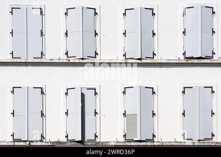 Eight windows with closed white shutters, one of them ajar, on the facade of an old white building Stock Photo