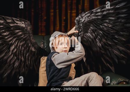 A boy with black angel wings sits in a dark room, playing on his phone. The boy looks bored and distracted, as if he is looking for something more in Stock Photo