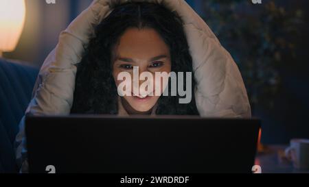 Smiling happy Hispanic female woman girl Indian Arabian lady smile using computer at late evening night cover under blanket typing on laptop watching Stock Photo
