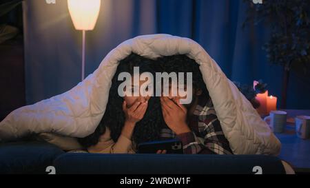 Couple Indian man and Arabian Hispanic woman night dating in bed under blanket family boyfriend girlfriend shocked watching horror film movie scared Stock Photo