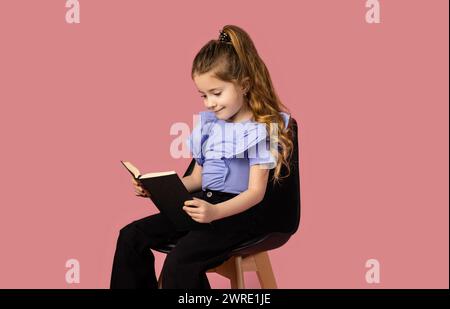 The portrait of a beautiful and stylish blonde girl, in a studio with a pink background, looks at the favorite book she is reading and smiles sweetly. Stock Photo
