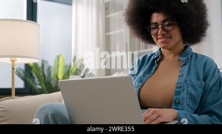 Busy African American woman using pc at home freelancer female user on couch sofa working with laptop online typing message e-learning student girl Stock Photo