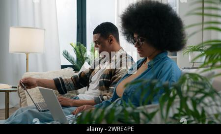 Busy workers freelancers African American man with woman couple at home couch browsing laptops computers pc devices online internet freelance work Stock Photo