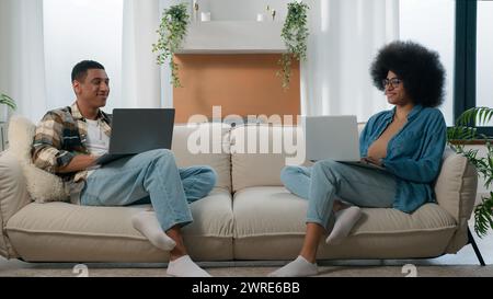 Happy couple relax at couch at home with laptop internet overuse freelancers African American man with woman smiling positive at sofa using typing Stock Photo