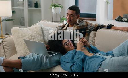 African American couple man with laptop computer sitting on couch home woman chatting smartphone browsing mobile phone laying on male knees girl guy Stock Photo
