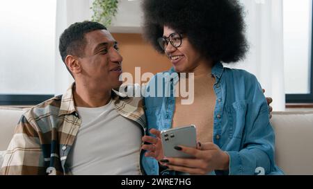 African American happy couple having fun using smartphone excited browsing mobile phone app at home girl and guy man and woman talking shopping Stock Photo