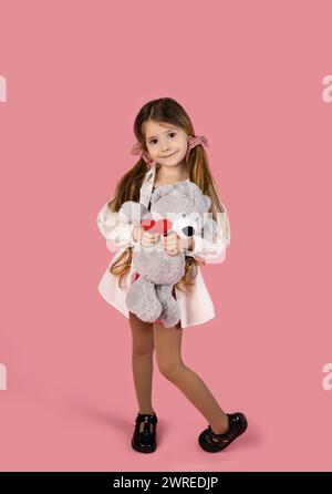 Full length portrait of a lovely little girl holding a teddy bear and a red paper heart, isolated on a pink background. High quality photo Stock Photo