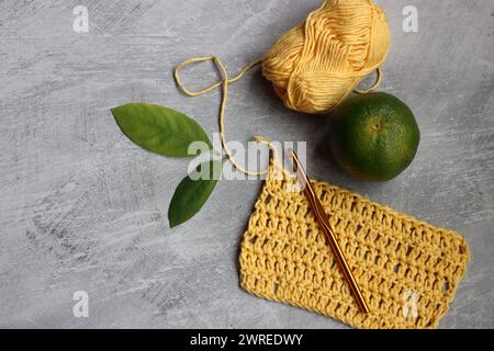 Crochet scarf in process. Crochet hook, yellow cotton yarn, lime and green leaves on grey background, top view Stock Photo