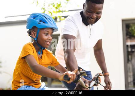 African American father teaches his son to ride a bike in the backyard, both smiling joyfully Stock Photo