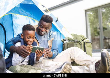 African American father and son share a moment with a smartphone inside a tent in their backyard Stock Photo