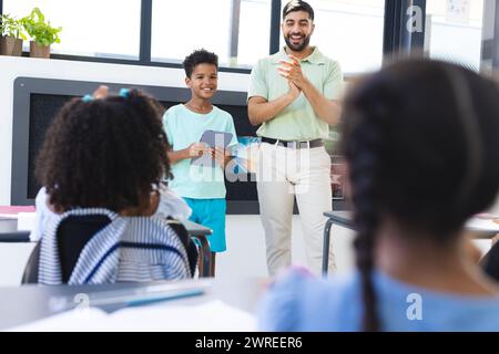 Young Asian male teacher stands applauding a biracial boy holding a tablet in a classroom in school Stock Photo