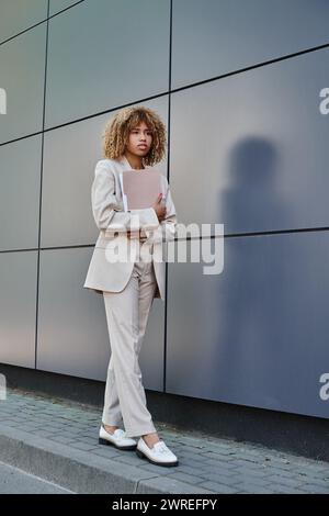 Confident young african american businesswoman in formal wear holding folder by grey office wall Stock Photo