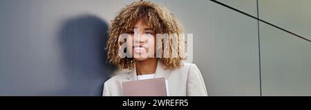 cheerful young african american businesswoman in formal wear holding folder by office wall, banner Stock Photo