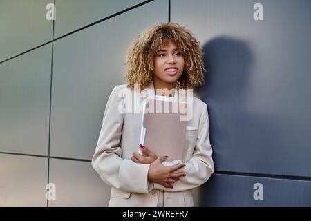 cheerful young african american businesswoman in formal wear holding folder by grey office wall Stock Photo