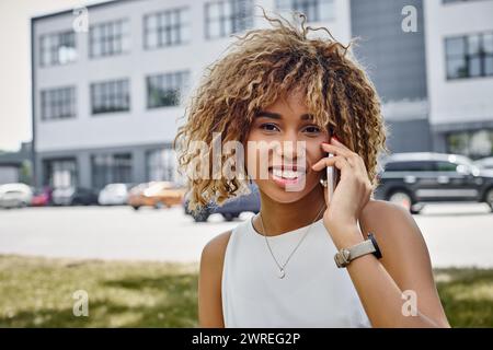 portrait of happy radiant curly-haired black woman having phone call in urban setting, smartphone Stock Photo