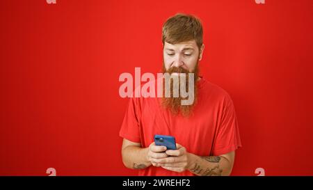 Focused redhead man engrossed texting on smartphone, serious face over isolated red background Stock Photo