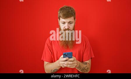 Focused redhead man engrossed texting on smartphone, serious face over isolated red background Stock Photo