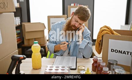 Charismatic young redhead guy, handsomely bearded, intently taking notes, addressing donations on his smartphone while sitting at a charity center tab Stock Photo