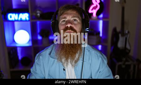 Staunch young redhead man streamer, seriously immersed in digital gaming world, donning headphones in the dimly lit gaming room Stock Photo