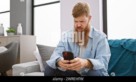 Focused young redhead man, chilling on his sofa indoors, engrossed in texting on his smartphone at his cozy apartment. Stock Photo