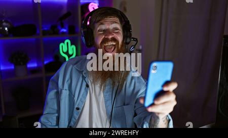 Grinning gaming wizard! young irish redhead man streamer confidently hosts epic video call via smartphone in heart of night-dark gaming room Stock Photo