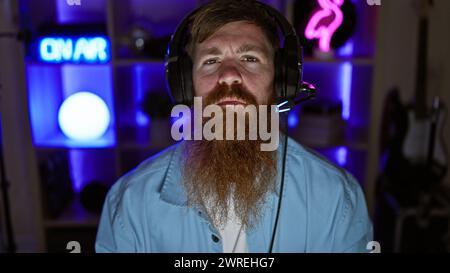 Staunch young redhead man streamer, seriously immersed in digital gaming world, donning headphones in the dimly lit gaming room Stock Photo