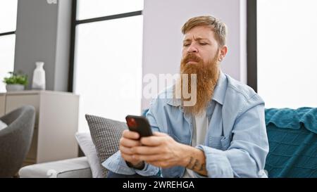 Focused young redhead man, chilling on his sofa indoors, engrossed in texting on his smartphone at his cozy apartment. Stock Photo