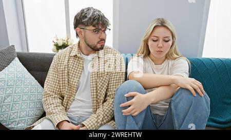 A man and woman sit apart on a couch, depicting tension or disagreement between them in a modern living room. Stock Photo