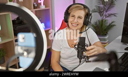 A smiling woman with headphones sits in a colorful gaming room at night in front of a computer and ring light. Stock Photo