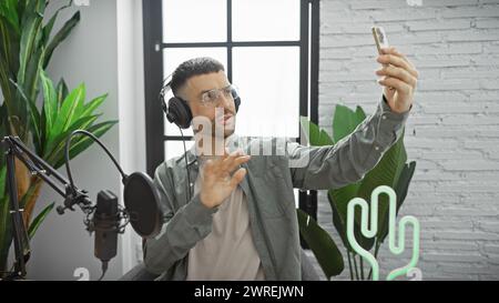 A young man engages in a lively podcast recording in a studio, where his headphones, microphone, and smartphone capture his enthusiasm. Stock Photo
