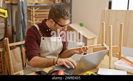 A bearded man carefully examines wooden material while using a laptop in his well-organized carpentry workshop. Stock Photo