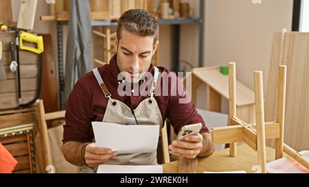 A focused hispanic man examines documents while holding a phone in a woodworking workshop. Stock Photo